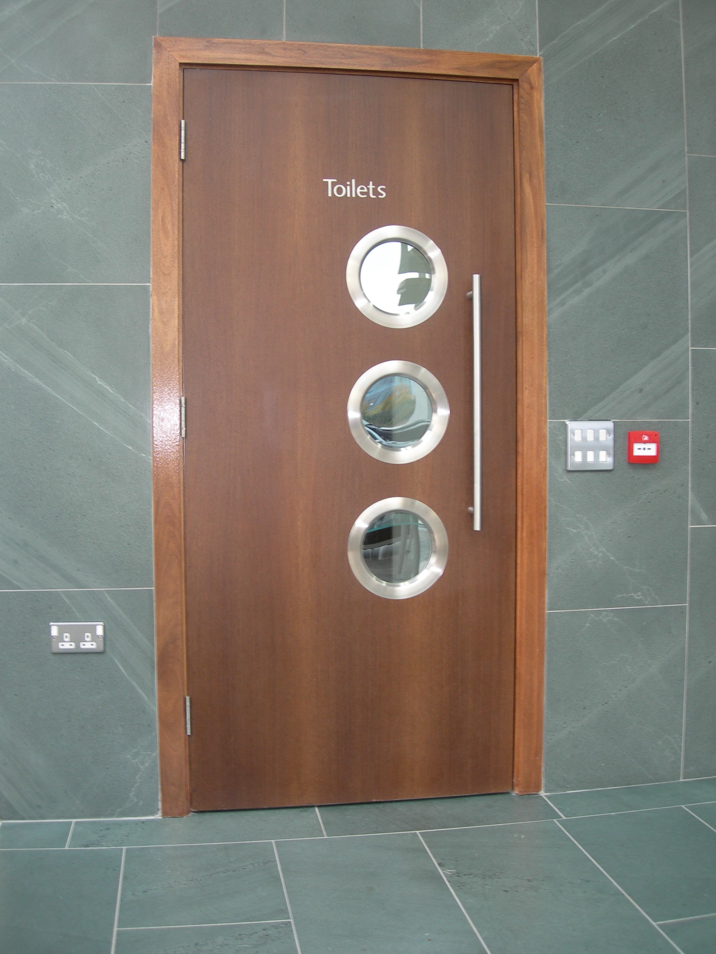 an image of a brown wooden door with three round silver slimport vision panels on a green tiled background